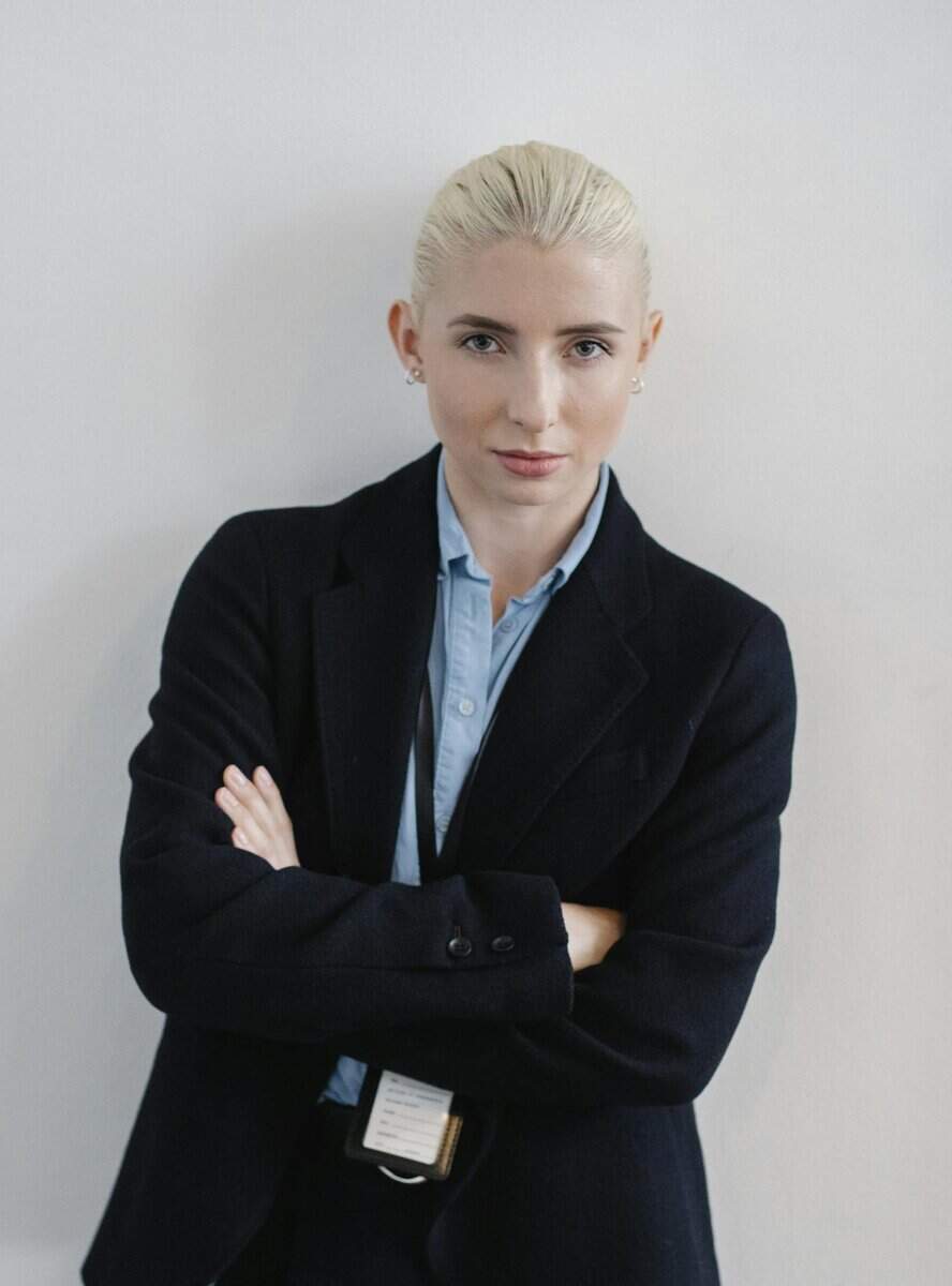 A person with light hair pulled back, wearing a dark blazer and light blue shirt, stands against a plain white background with arms crossed.