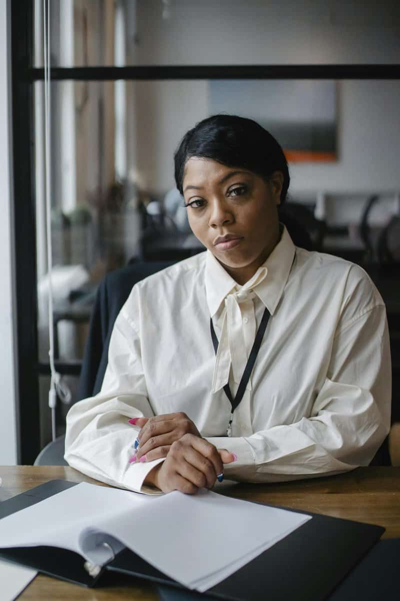 A woman in a white blouse sits at a desk with an open folder and papers, looking directly at the camera in an office setting.