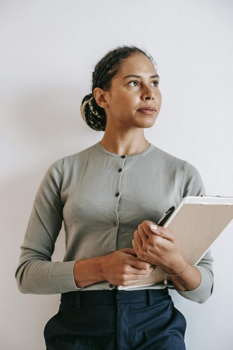 A woman wearing a light green cardigan holds a clipboard and pen while looking to the side against a plain light background.