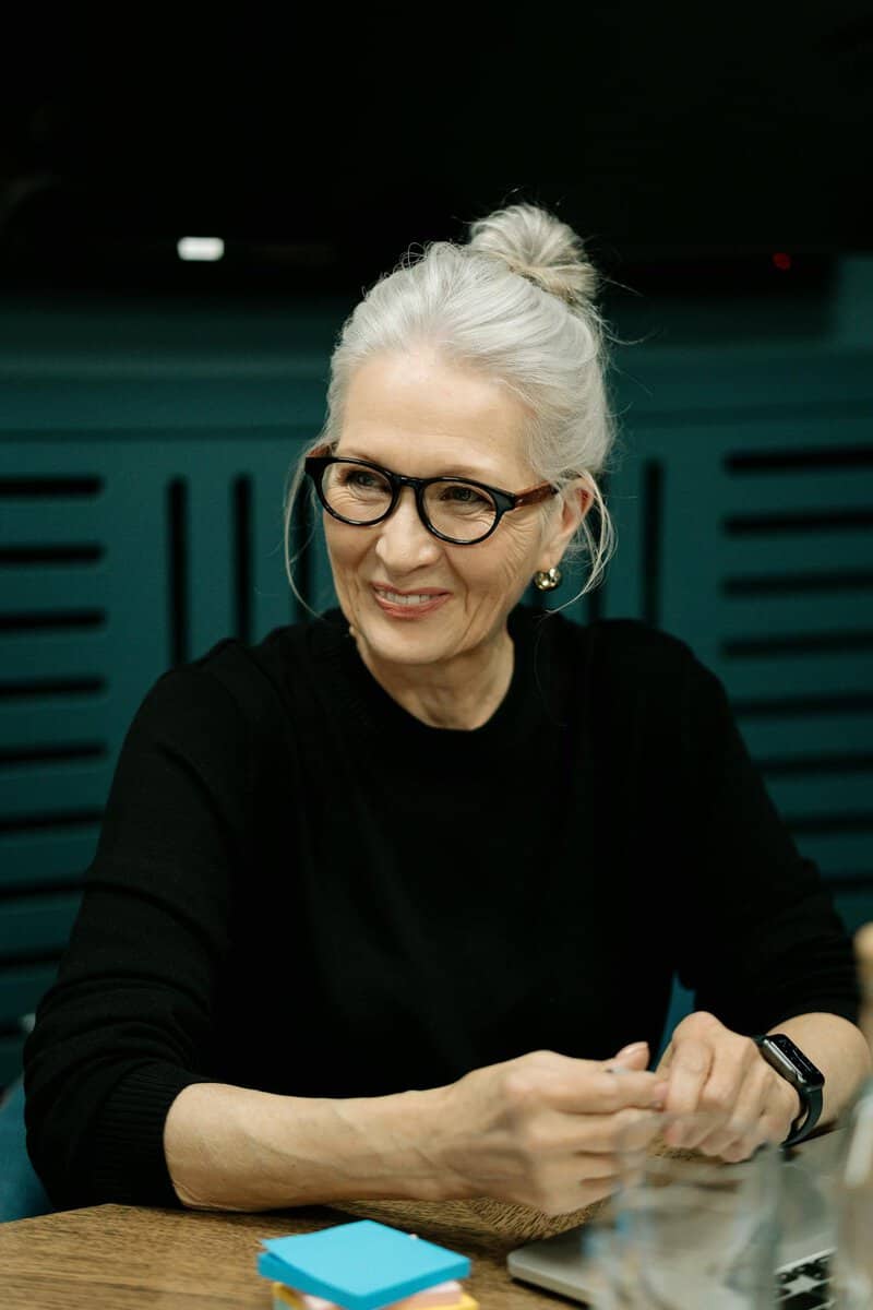 An older woman with gray hair in a bun, wearing glasses and a black top, sits at a table with a small stack of blue sticky notes in front of her.