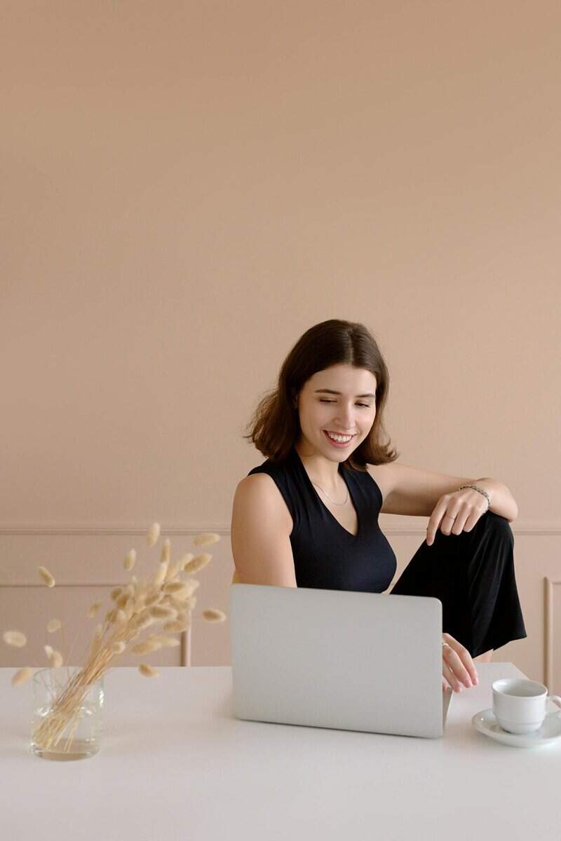 A woman sits at a white table, smiling while looking at a laptop. A cup and saucer are nearby, along with a vase of dried grasses.